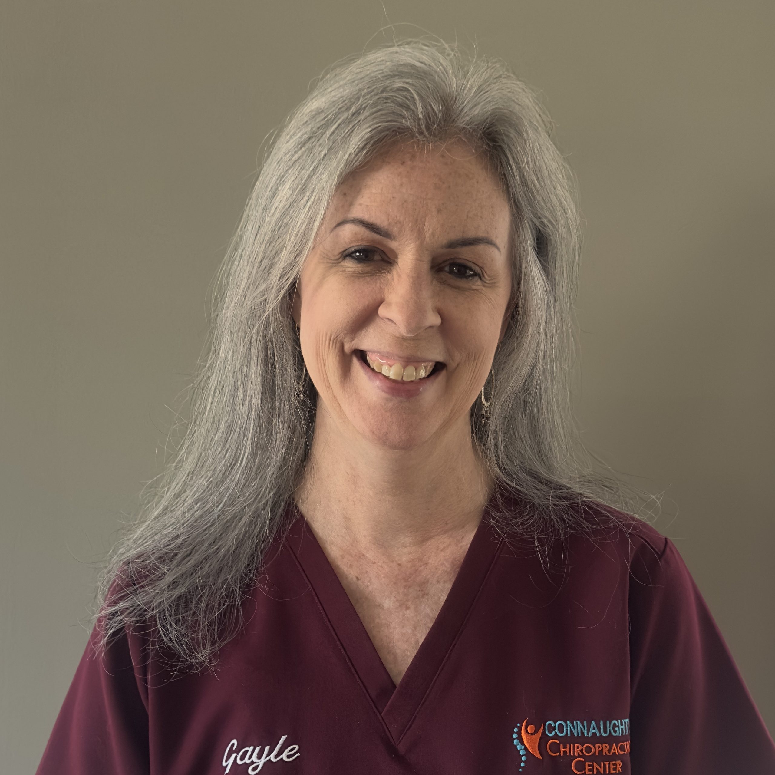 Chiropractic Old Saybrook CT Gayle Gayle standing in front of a tan wall in maroon scrubs smiling